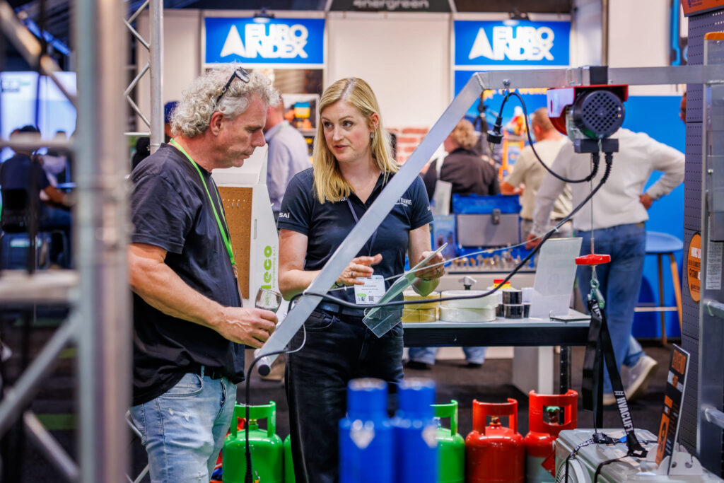 Standhouder in gesprek met een bezoeker tussen gasflessen en installatiemateriaal